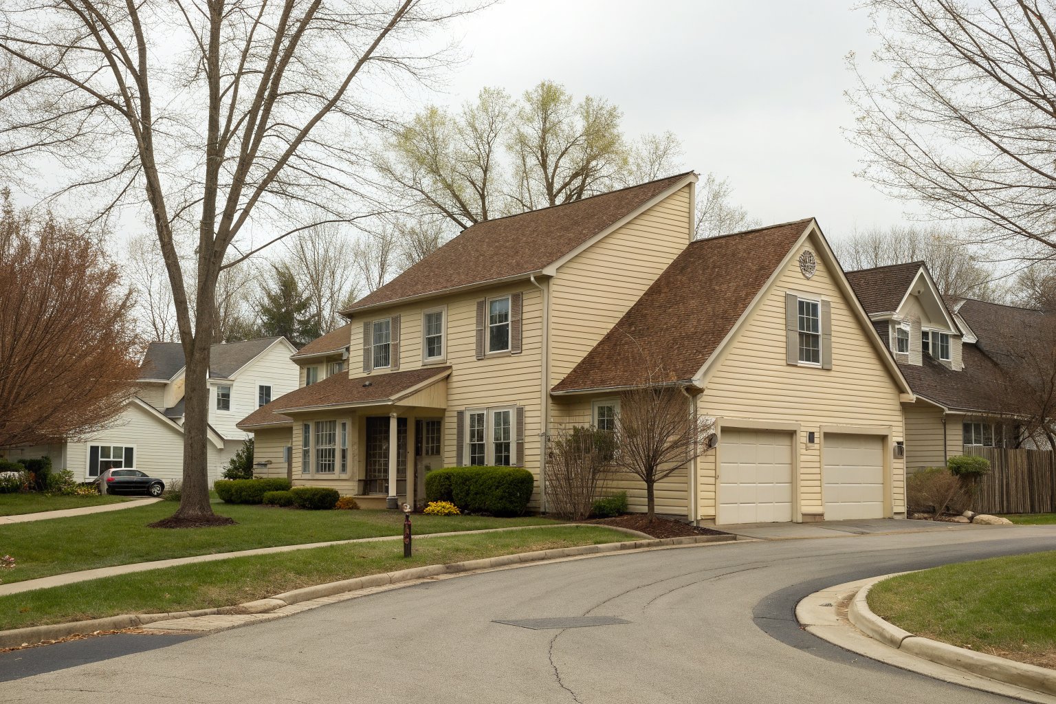 A two-story colonial house with tan siding and a brown shingle roof on a curving street